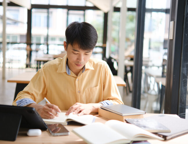 A student engaged in online learning, taking notes and reviewing study materials with books and digital devices to prepare for exams.